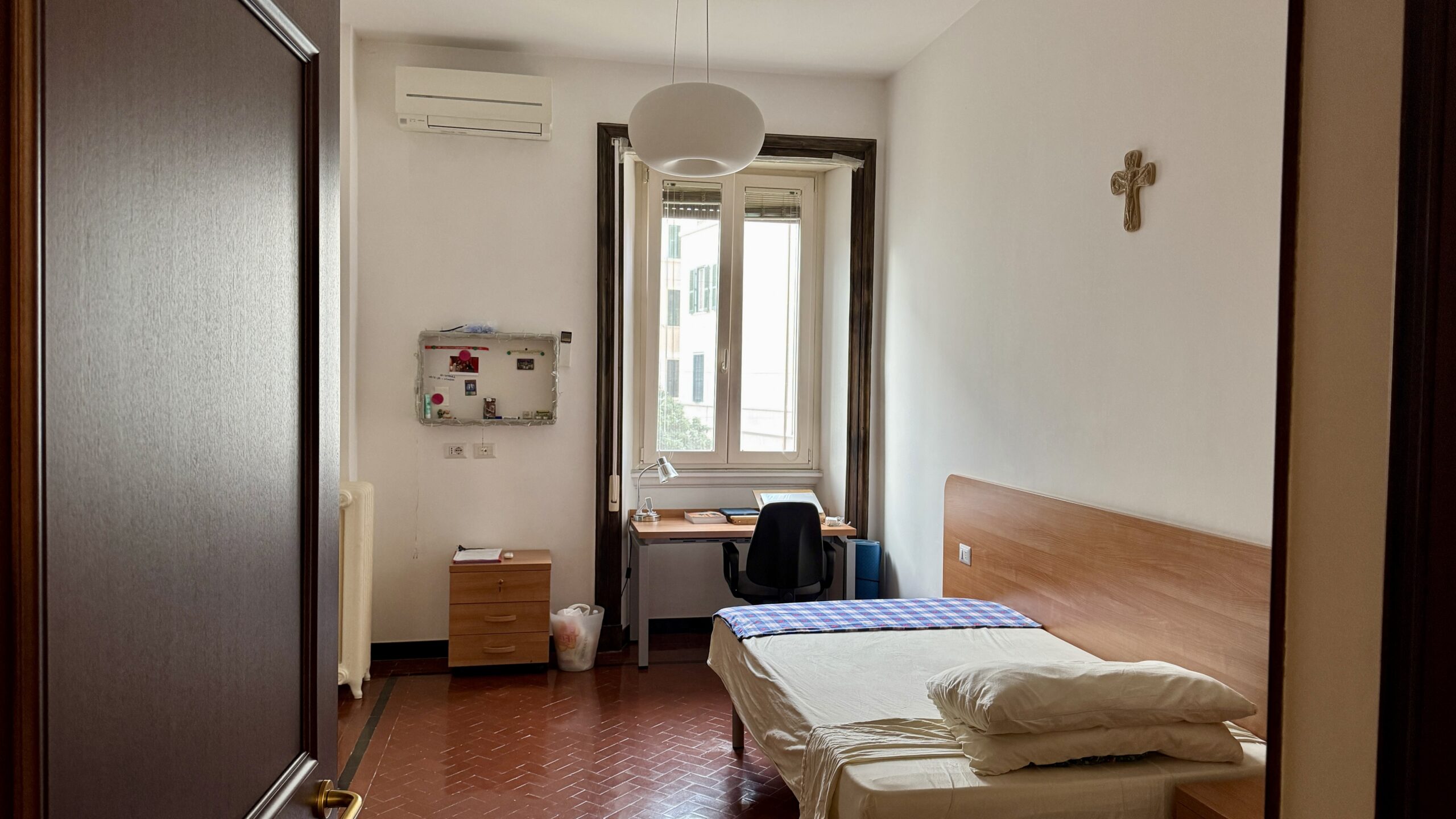 Interior de habitación individual, con cama, escritorio de frente a la ventana, aire acondicionado y calefactor.